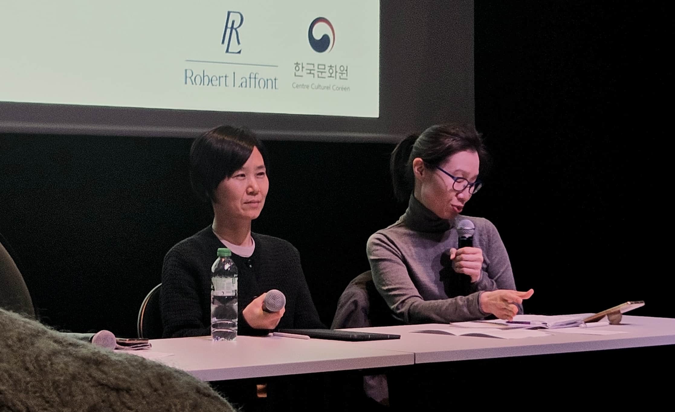 Cho Nam-Joo et son interprète assises à une table blanche lors d'une conférence, chacune tenant un microphone. En arrière-plan, une projection affichant les logos de l'éditeur Robert Laffont et du Centre Culturel Corée. Des documents et une bouteille d'eau sont posés sur la table.