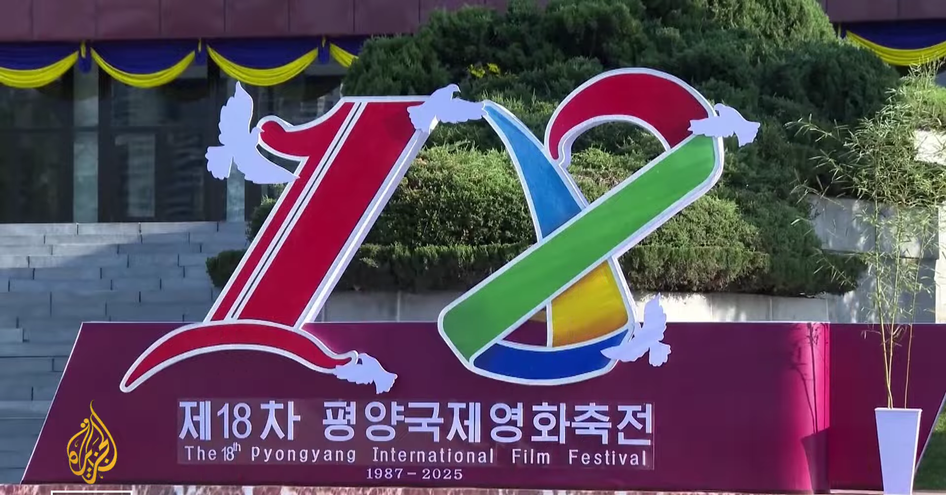 Panneau officiel de la 18e édition du Pyongyang International Film Festival, affichant le logo coloré de l'événement avec le chiffre 18 avec des colombes blanches sur un fond bordeaux avec les inscriptions en coréen et en anglais mentionnant les dates de 1987-2025.