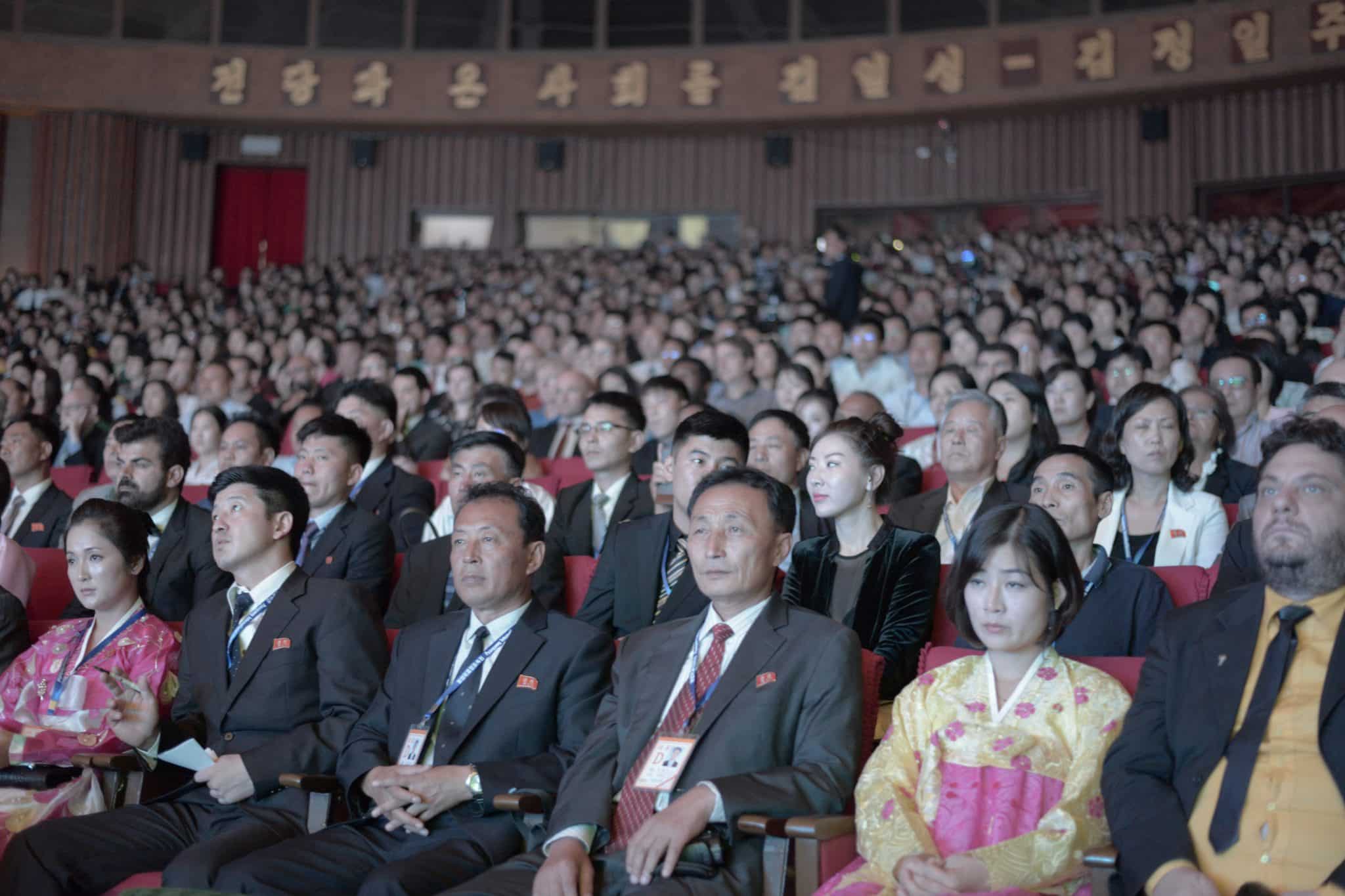 Salle de projection du Pyongyang International Film Festival remplie d'un public nombreux, mêlant spectateurs nord-coréens en costume officiel et traditionnel et visiteurs étrangers, certains portant des accréditations, dans un grand auditorium aux sièges rouges.