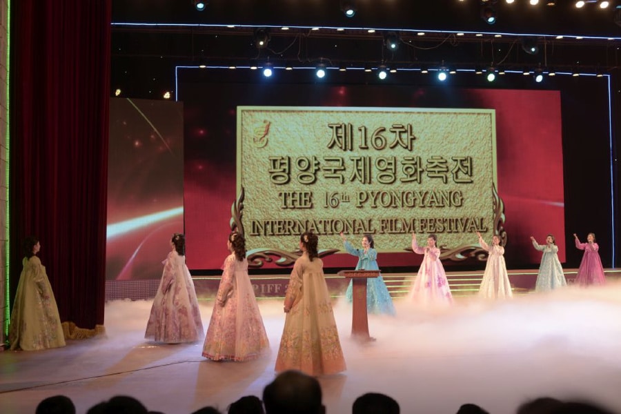 Cérémonie d'ouverture de la 16e édition du Pyongyang International Film Festival, avec plusieurs femmes en hanbok traditionnel coloré sur scène, devant un grand écrant affichant le nom du festival en coréen et en anglais, dans une salle de spectacle illuminé dans la capitale de la Corée du Nord à Pyongyang.