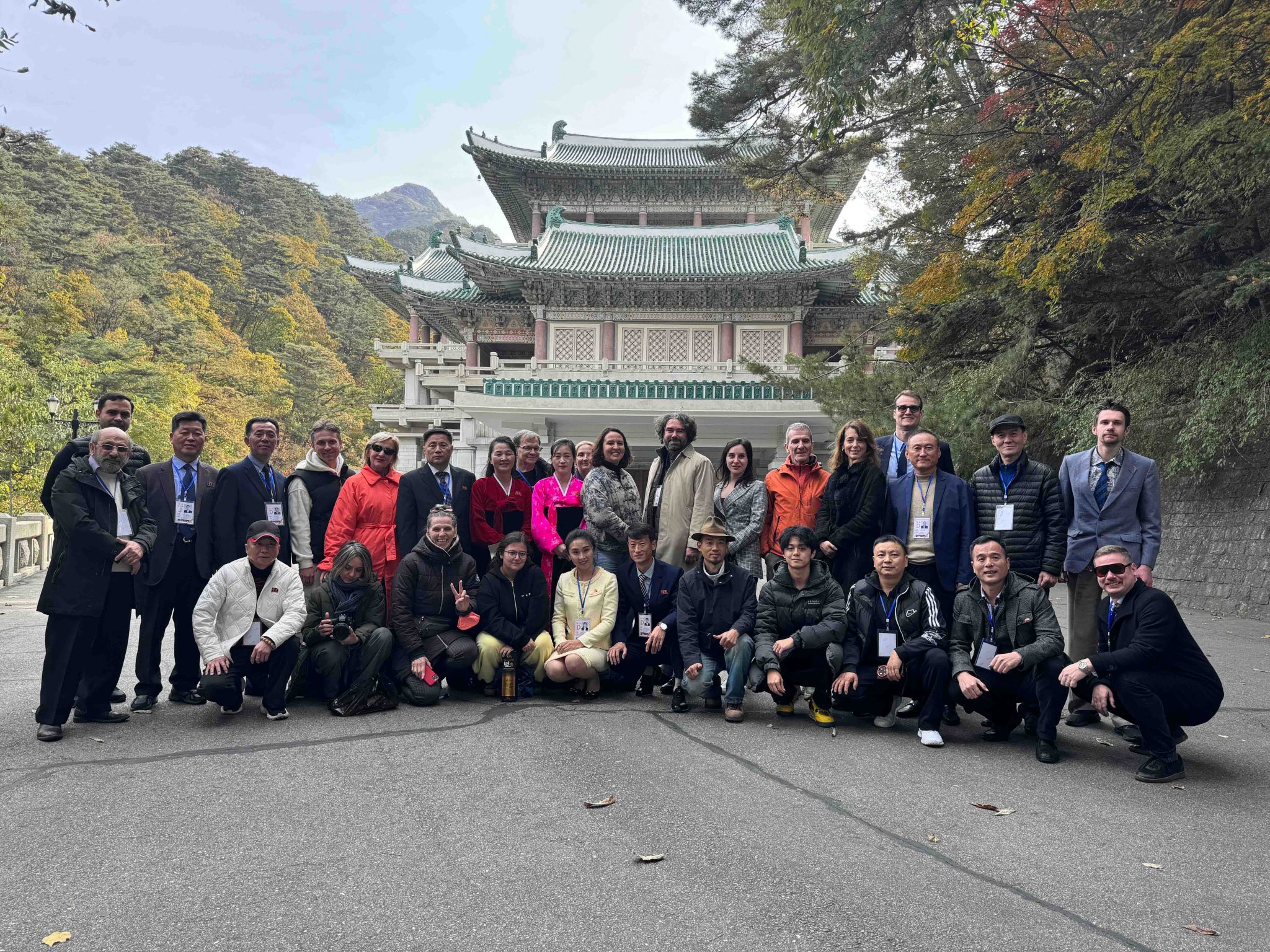 Photo de groupe d'une délégation étrangère de la 18e édition du Pyongyang International Film Festival, posant devant un édifice traditionnel coréen aux toits verts en automne, avec des participants portant des accréditations et des guides nord-coréens reconnaissables à leurs badges officiels.