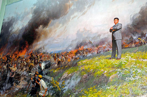 Peinture propagandiste nord-coréenne représentant Kim Jong-il debout, bras croisés, dominant en surplomb une scène de tournage cinématographique épique avec des explosions, des flammes et une équipe de tournage en action au premier plan.