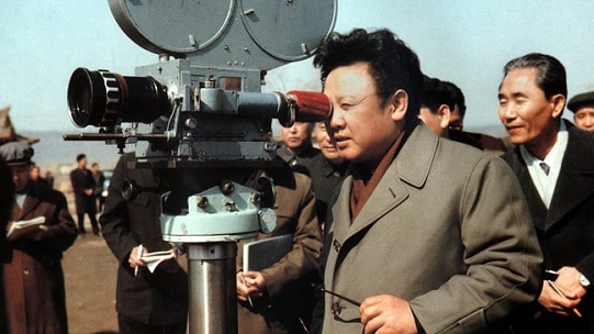 Photographie en couleur de Kim Jong-il regardant à travers l'objectif d'une caméra de cinéma sur un tournage extérieur, entouré de plusieurs personnes, en mars 1979.
