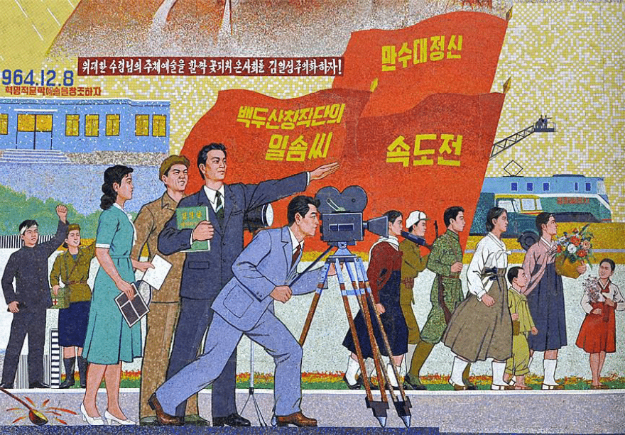 Mosaïque nord-coréenne représentant une scène de tournage cinématographique, avec un caméraman au premier plan, entouré de personnages en tenue populaire et militaire, sur fond de drapeaux rouges et de slogans en coréen