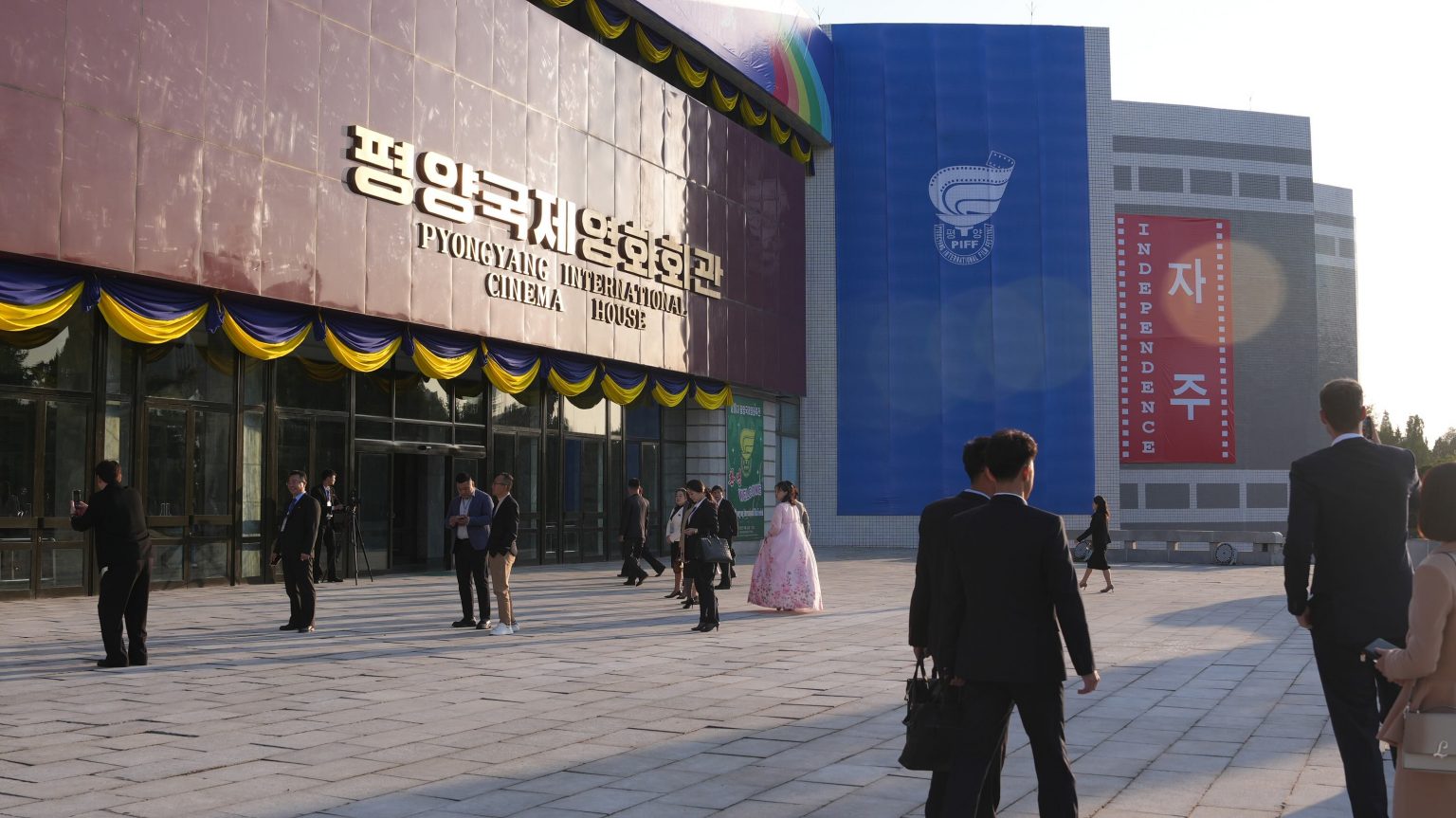 Façade du Pyongyang International Cinema House, salle principale du Pyongyang International Film Festival (PIFF), décorée de guirlandes bleues et jaunes, avec des visiteurs en tenues formelles et traditionnelles devant l'entrée et une grande bannière bleue affichant le logo du festival et le mot "independance" en anglais et en coréen