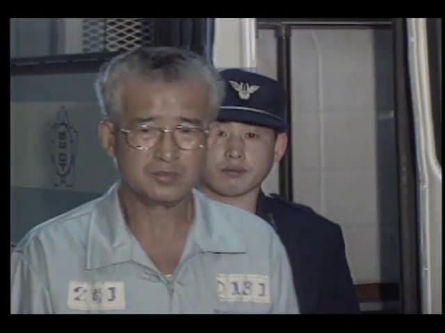 Jeong Su-il portant des lunettes et une tenue de détenu grise avec des numéros d'écrou, escorté par un agent pénitentiaire en uniforme bleu marine.