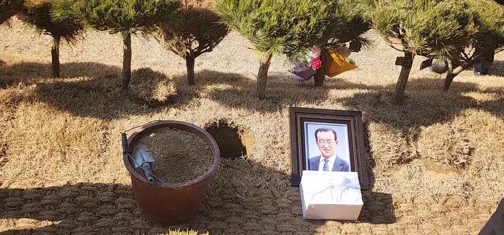 Site d'inhumation en plein air entouré de pins, avec une urne funéraire en terre cuite, un portrait encadré de Jeong Su-il, Muhammad Kansu en costume sombre, et un paquet blanc noué d'un ruban, posés sur un sol recouvert d'herbe sèche. Des fleurs sont visibles en arrière-plan.