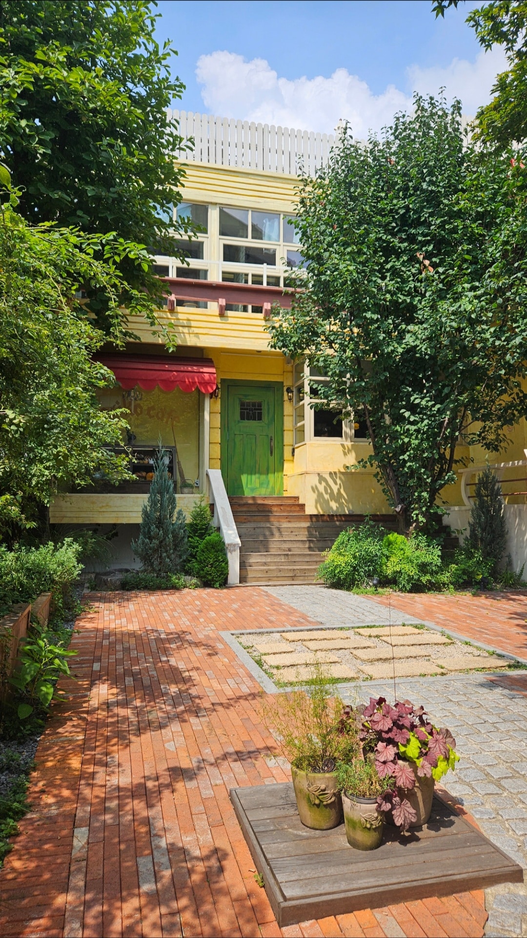 Façade colorée jaune et verte d'un café coréen entouré d'arbres et d'un jardin verdoyant, avec une allée en briques menant à l'entrée. C'est le Koriko Café en Corée du Sud