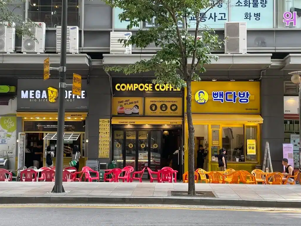 Trois cafés côte côte dans une rue en Corée du Sud : Mega Coffee, Compose Coffee et Paik's Coffee (백다방), avec des chaises colorées disposées sur le trottoir.