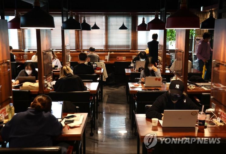 Un café coréen avec des tables en bois qui sont alignées, entièrement occupées par des personnes travaillant et étudiants sur leurs ordinateurs portables, illustrant le phénomène des Cagongjok (카공족)