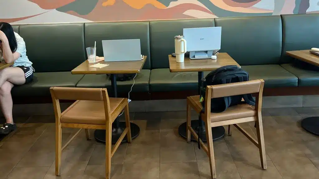 Deux ordinateurs portables ouverts sur des tables individuelles dans un café coréen, avec un sac à dos posé sur une chaise.
