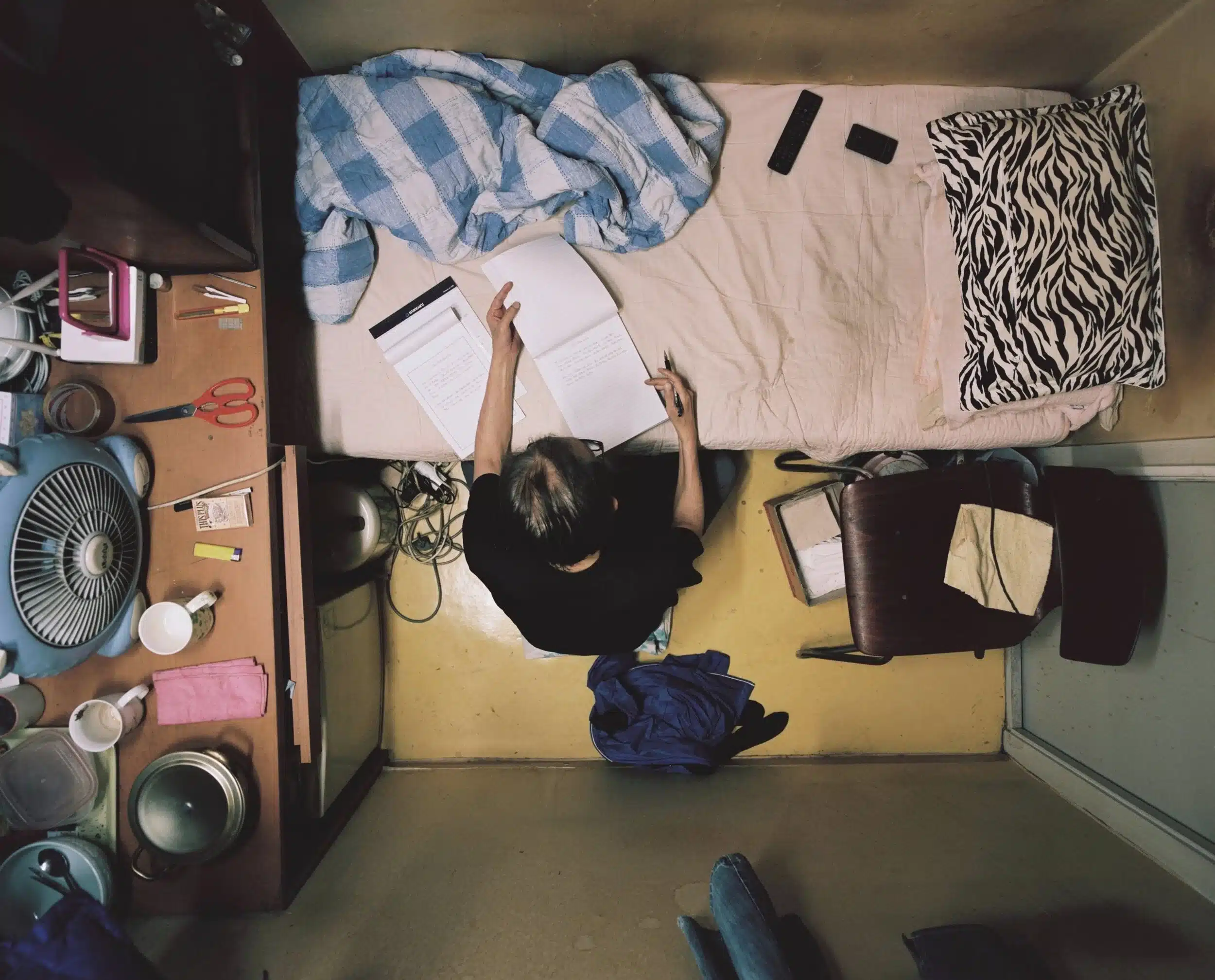 Vue plan de haut d'un Goshiwon (고시원) de Corée du Sud. Une personne étudie, travaille, assise par terre dans une petite chambre, entourée d'un lit simple, d'une table encombrée et de quelques affaires personnelles. Goshiwon (고시원).